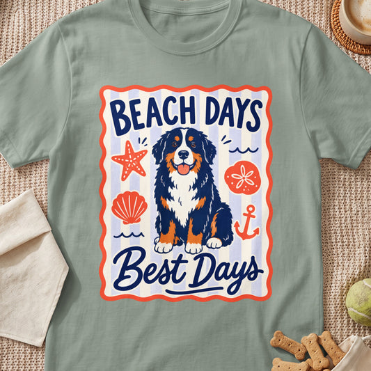 A sage green t-shirt features a graphic of a Bernese Mountain Dog surrounded by beach elements and text.