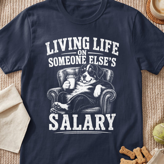 A dark blue t-shirt printed with a Bernese Mountain Dog wearing sunglasses and holding a coffee cup while lounging in a leather armchair, accompanied by the text "LIVING LIFE ON SOMEONE ELSE'S SALARY".