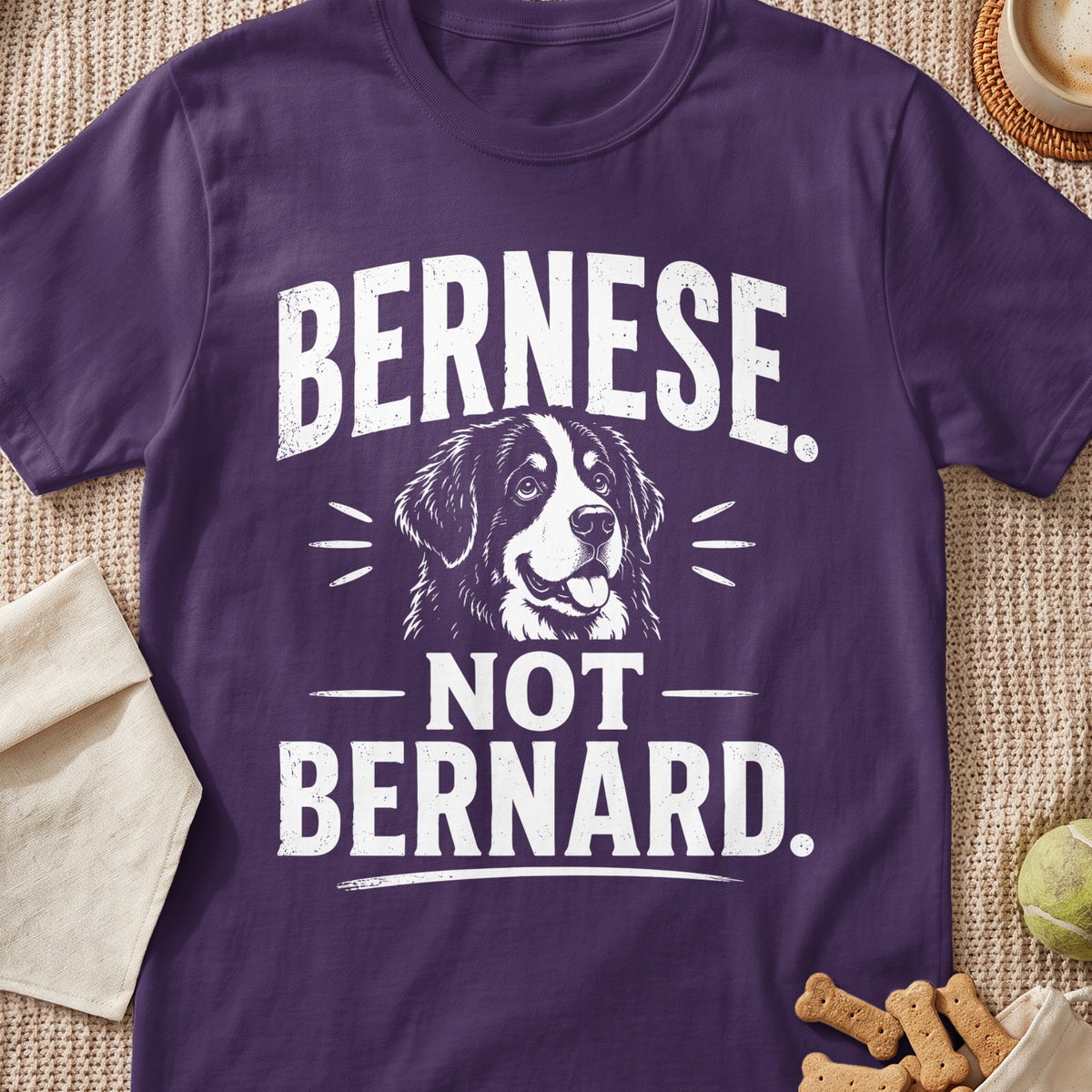 A purple t-shirt featuring white graphic text that reads Bernese Not Bernard surrounding a Bernese Mountain Dog illustration.