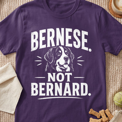 A purple t-shirt featuring white graphic text that reads Bernese Not Bernard surrounding a Bernese Mountain Dog illustration.