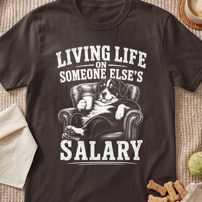 A dark brown t-shirt featuring a graphic of a Bernese Mountain Dog relaxing in an armchair with sunglasses and a coffee cup, accompanied by the text "Living Life on Someone Else's Salary."