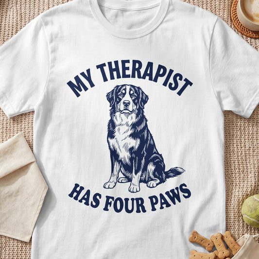 White t-shirt with dark blue text "MY THERAPIST HAS FOUR PAWS" and dog illustration.