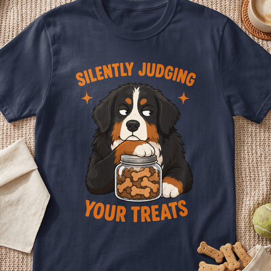 A dark blue t-shirt with an illustration of a Bernese Mountain Dog resting its chin on a jar of dog treats, accompanied by the text SILENTLY JUDGING YOUR TREATS.