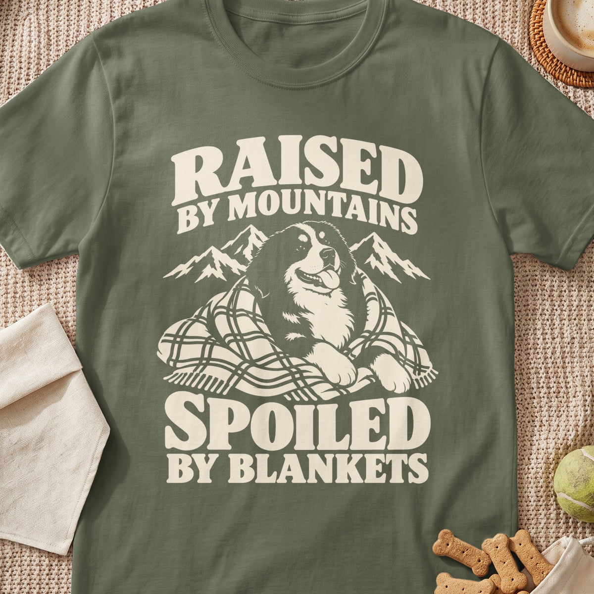 Olive green t-shirt with dog, mountains, and text "Raised by Mountains, Spoiled by Blankets."