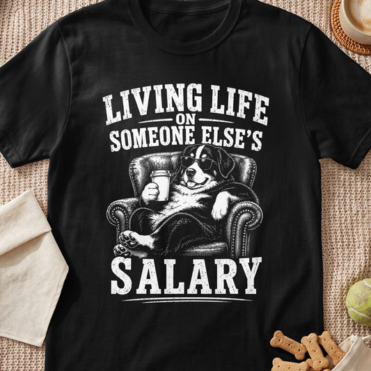 A black t-shirt featuring a graphic of a Bernese Mountain Dog wearing sunglasses, lounging in an armchair, holding a coffee cup, and the text LIVING LIFE ON SOMEONE ELSE’S SALARY.