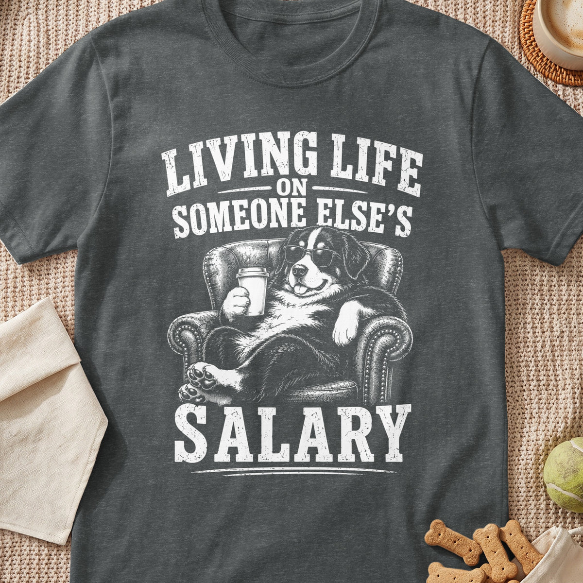 Dark gray t-shirt with a graphic of a dog in sunglasses relaxing in an armchair holding a coffee cup, and the text Living life on someone else's salary.
