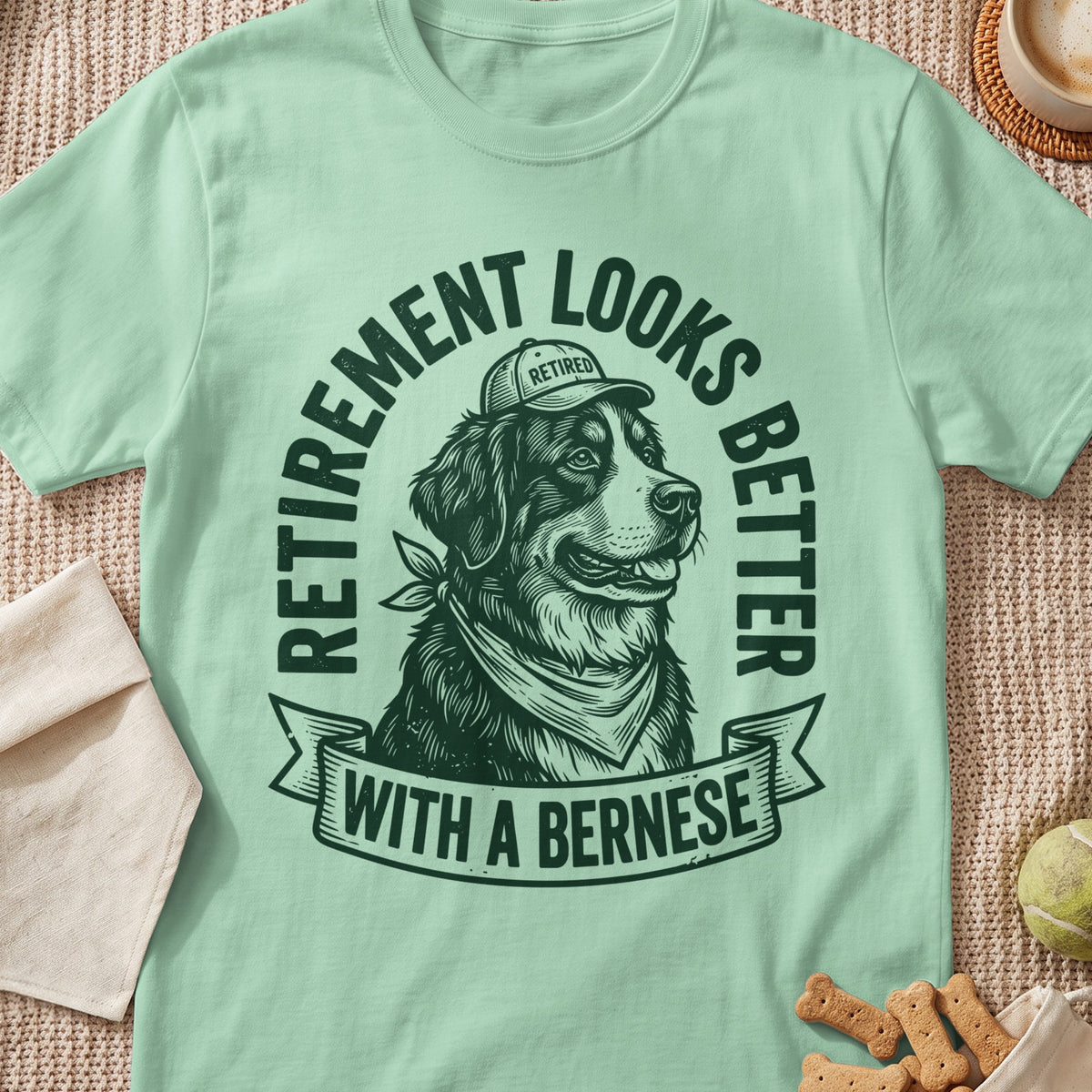 A light green t-shirt featuring a graphic of a Bernese Mountain Dog wearing a hat and bandana with text reading Retirement looks better with a Bernese.