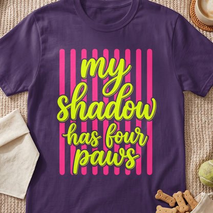 Purple t-shirt with neon green text reading "my shadow has four paws" on pink stripes.